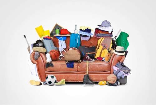 A cluttered sofa with a variety of discarded items, including clothing, shoes, sports equipment, and small household objects, all piled on and around the armrests and seat cushions. The fabric of the sofa is upholstered in a textured, reddish-brown material. Among the objects are a black and white soccer ball, a basketball, a beige handbag, a pair of sneakers, a yellow safety helmet, and a baseball cap. The items also include a tennis racket, a stack of folded clothes, a small whiteboard, and plastic containers, some of which are in bright colours like yellow and green. The scene appears to be set outdoors, possibly on a driveway or pavement, with light neutral background suggesting an environment ready for rubbish removal or clearance. Victoria House Clearance, a waste management company, may typically handle such clutter as part of their house clearance services in Victoria, South London, or surrounding areas. The overall visual indicates a need for professional rubbish collection and removal to clear the space efficiently and safely.