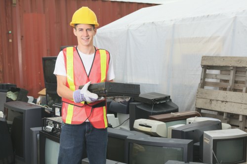 A young man wearing a yellow safety helmet, high-visibility vest with pink and yellow reflective stripes, and light grey gloves is standing outdoors in front of an array of discarded electronic waste, including old televisions and computer monitors, placed on a paved surface. Behind him, there is a red wooden fence and a white plastic tent or cover, suggesting a waste collection or clearance area. The man is smiling and holding a black electronic device with both hands. The surrounding environment hints at an organized rubbish removal operation, relevant to services offered by Victoria House Clearance in Victoria, with the focus on proper disposal of electronic waste. The setting appears to be daytime with natural lighting illuminating the scene, emphasizing the materials and textures of the electronic items and safety equipment.