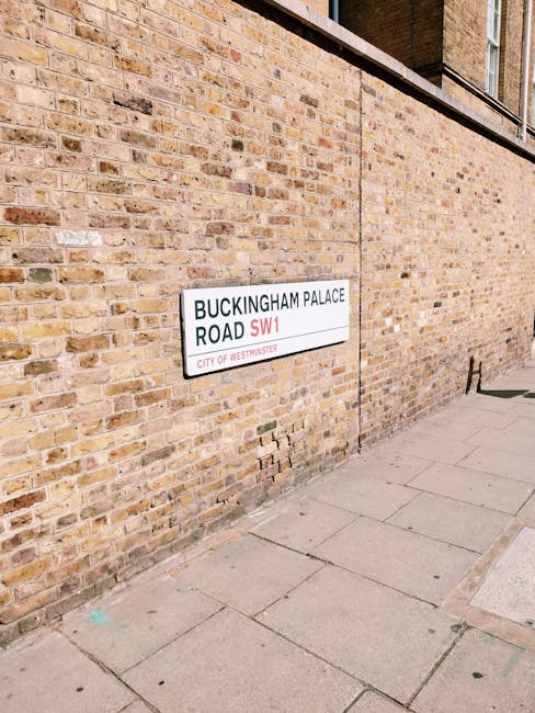 A rectangular street sign is mounted on a brick wall adjacent to a paved sidewalk on Buckingham Palace Road in Westminster. The sign displays black text on a white background, reading 'BUCKINGHAM PALA