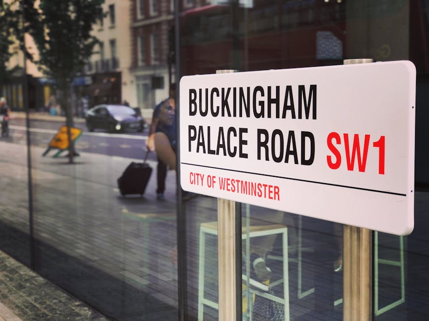 A rectangular street sign is mounted on a brick wall adjacent to a paved sidewalk on Buckingham Palace Road in Westminster. The sign displays black text on a white background, reading 'BUCKINGHAM PALA