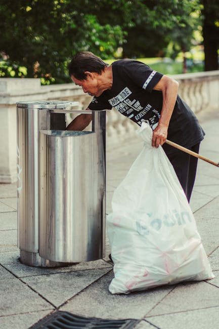 A sanitation worker dressed in a blue uniform with an orange high-visibility vest and matching blue cap is emptying a blue wheeled bin into a white waste collection truck on a cobbled urban street. Th